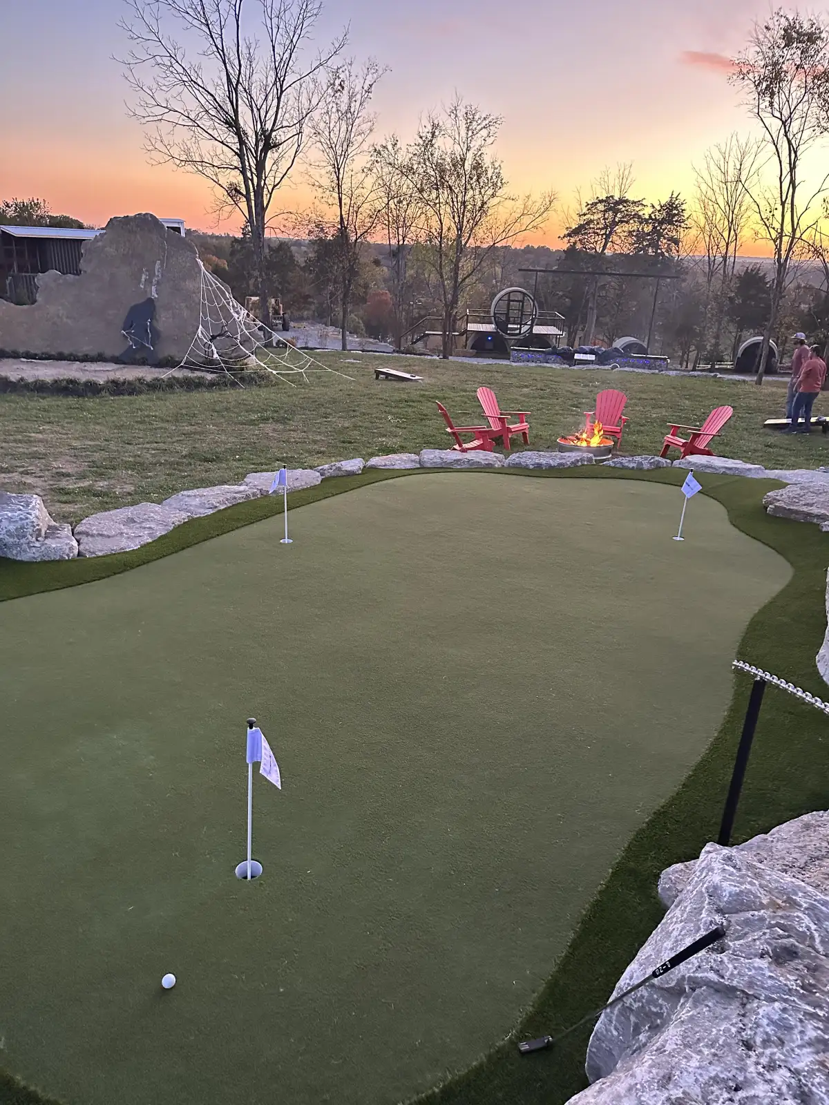 Stunning sunset view of a contoured backyard putting green with flags, fire pit, and lounge chairs in Michigan